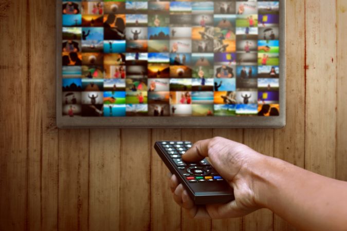 Fernbedienung in der Hand eines Mannes zeigt auf Fernseher mit zahlreichen unterschiedlichen Kacheln unterschiedlicher Filmangebote