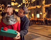 Mann hält Frau von hinten die Augen zu und gibt ihr ein Geschenk