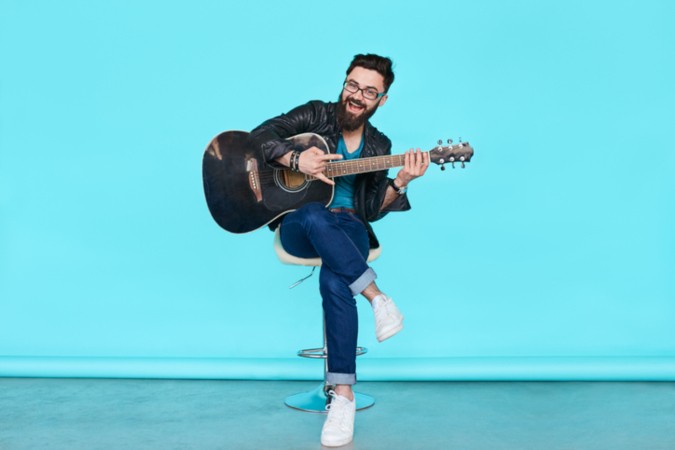 Mann mit Gitarre sitzt auf Stuhl vor blauem Hintergrund