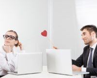 Mann und Frau sitzen vor Laptops. Er hält einen Herz-Lolli und guckt verliebt.